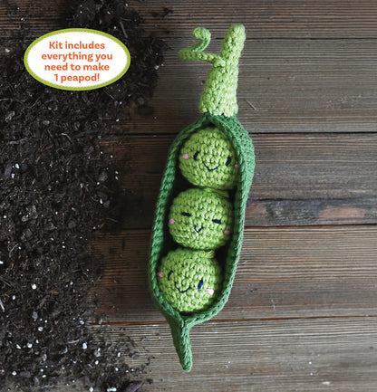 Crocheted green peas in a pod with smiling faces on a wooden surface next to soil.