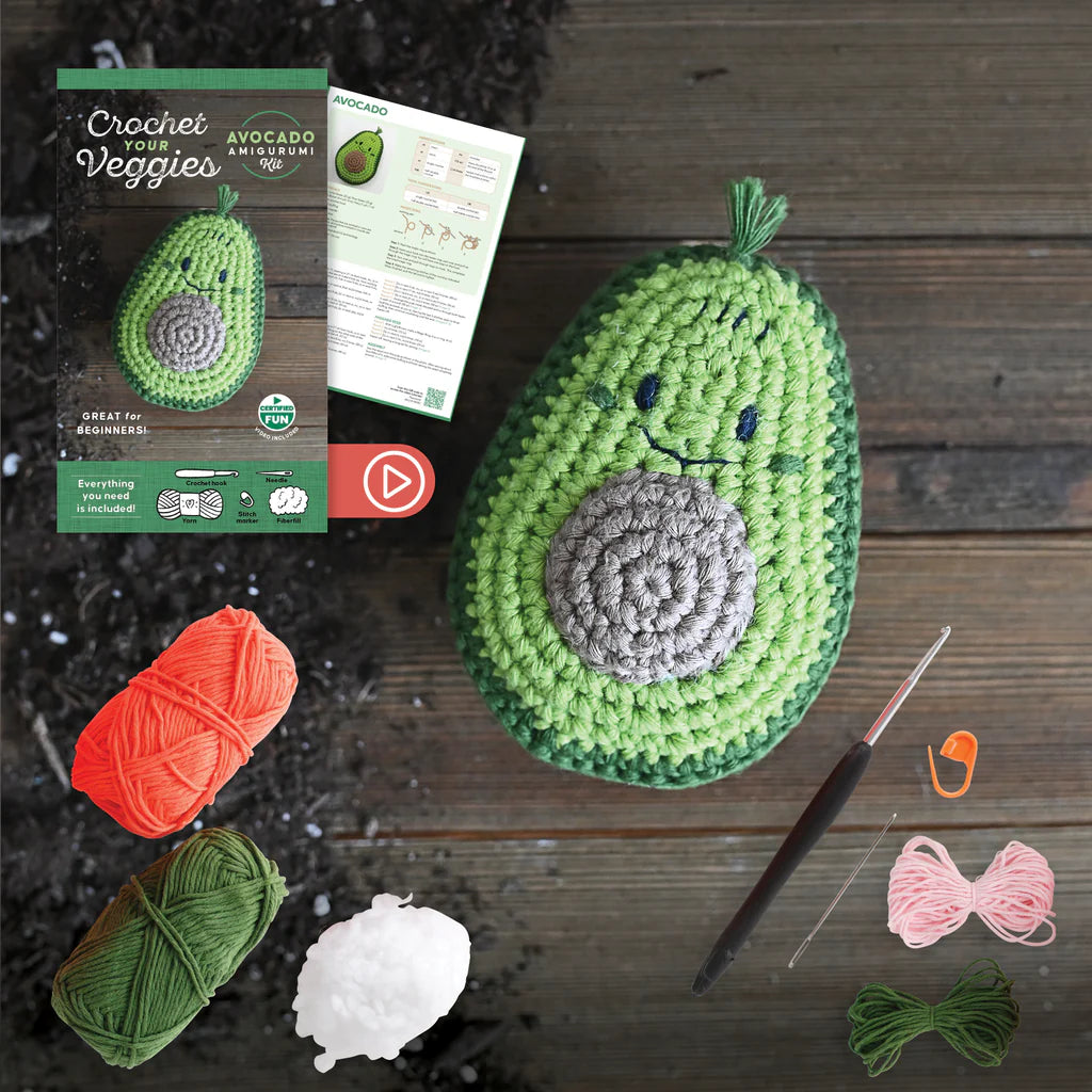 Crocheted avocado with yarn, hooks, and a pattern leaflet on a wooden surface.