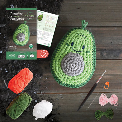 Crocheted avocado with yarn, hooks, and a pattern leaflet on a wooden surface.