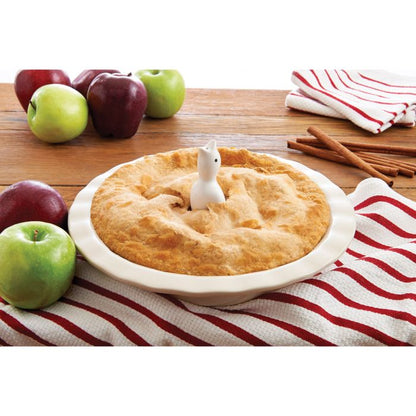 Apple pie with a pie bird on top, surrounded by apples and cinnamon sticks on a wooden table.