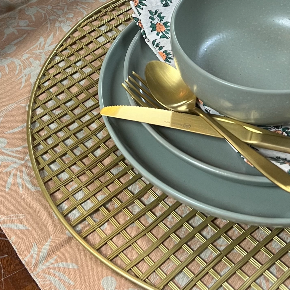angled view of gold wire charger with a sage green place setting and gold silverware on it set on a blush floral runner.
