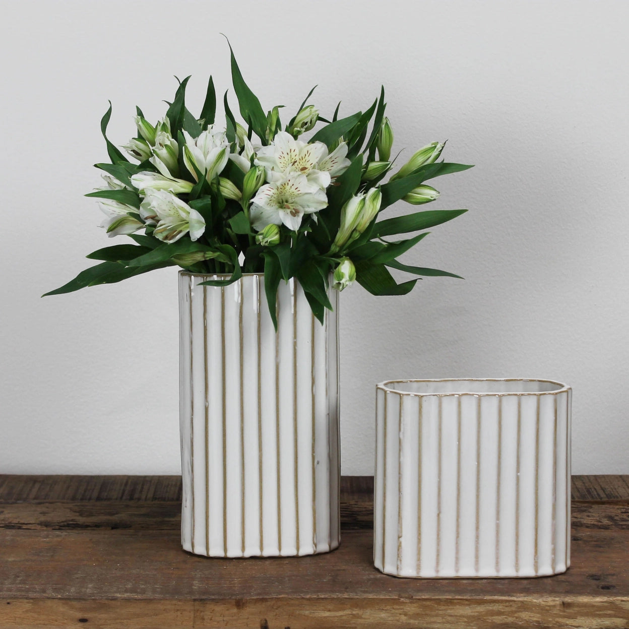 Two white ceramic vases with vertical stripes on a wooden surface against a plain background, larger one has flowers in it