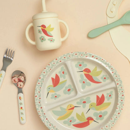 hummingbird plate, cup, and silverware arranged on a pale yellow background.