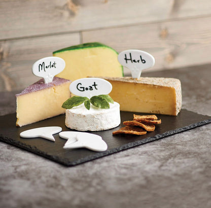 3 cheese markers labeled with either merlot, goat, or herb in a block of cheese arranged on a stone slab with crackers and herbs.