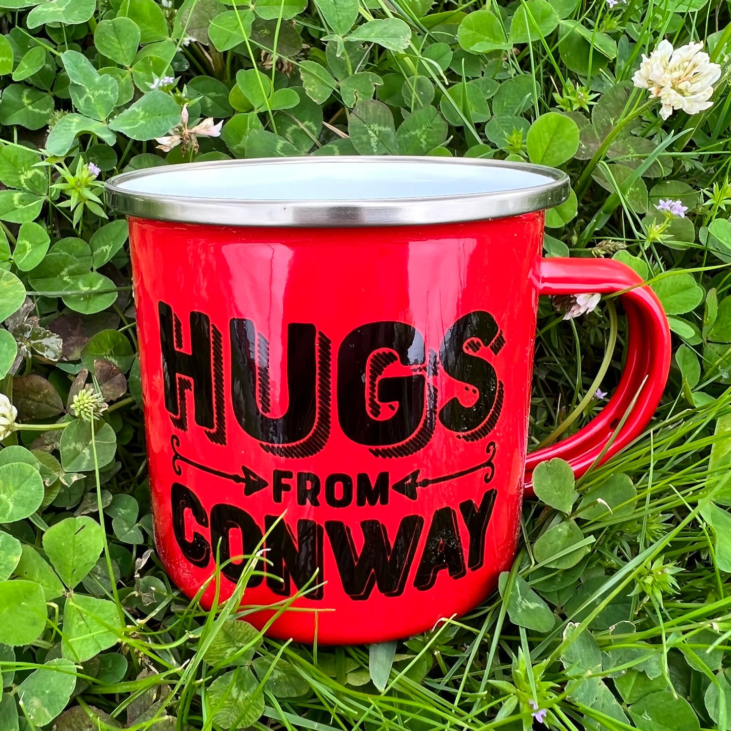 red tin mug with "hugs from conway" in black laying in a field of clovers.