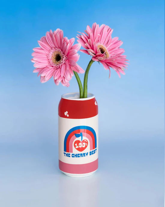 white vase shaped like a can of soda with red, pink, and blue design the reads " 100% the cherry best" and has 2 pink daisies in it.