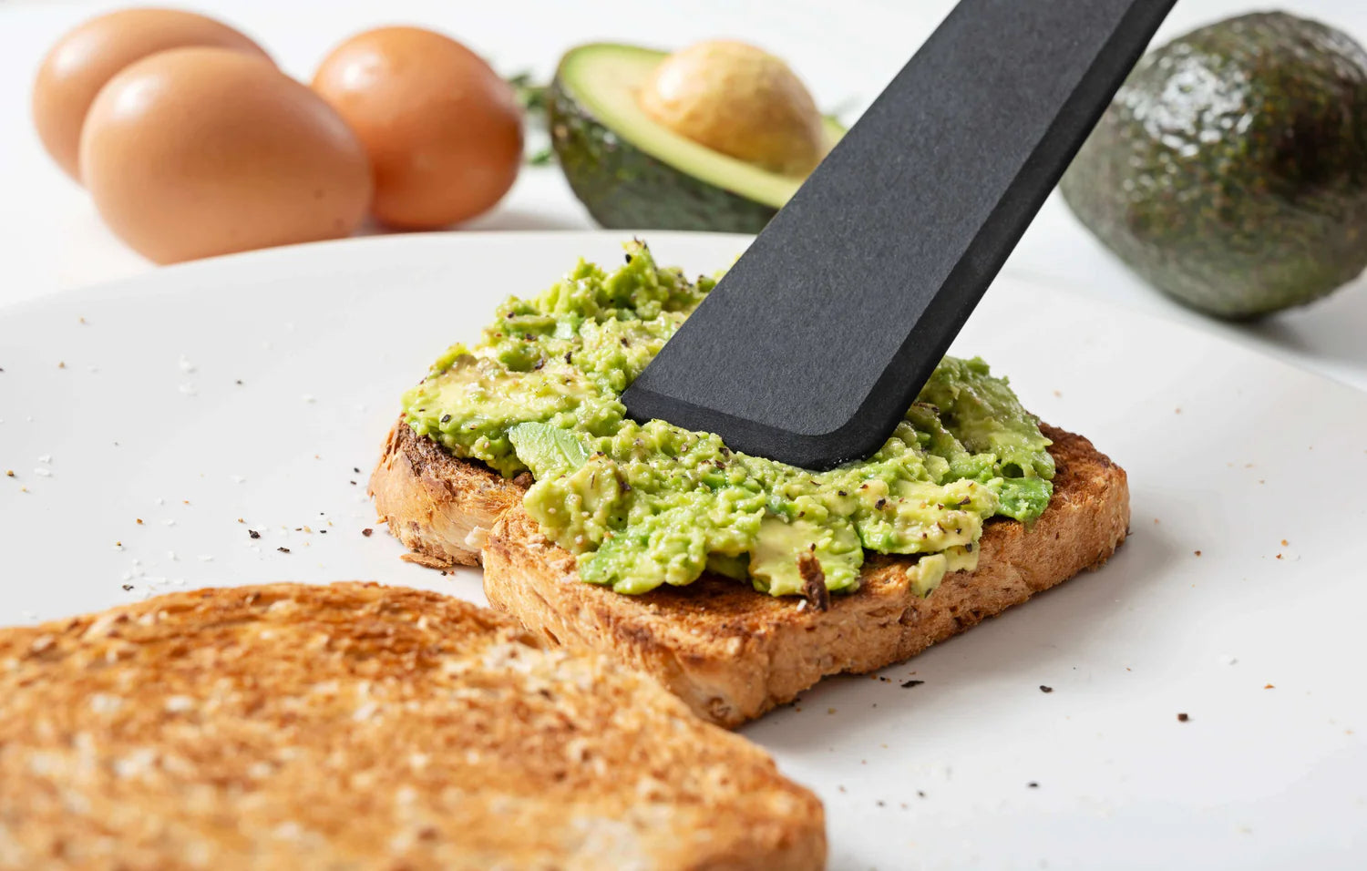 Avocado spread on toast with a black spatula, surrounded by avocados and eggs on a white surface.