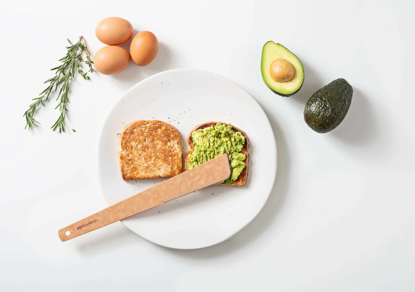 Avocado toast on a white plate with wooden utensil and avocados, eggs, rosemary arranged around it