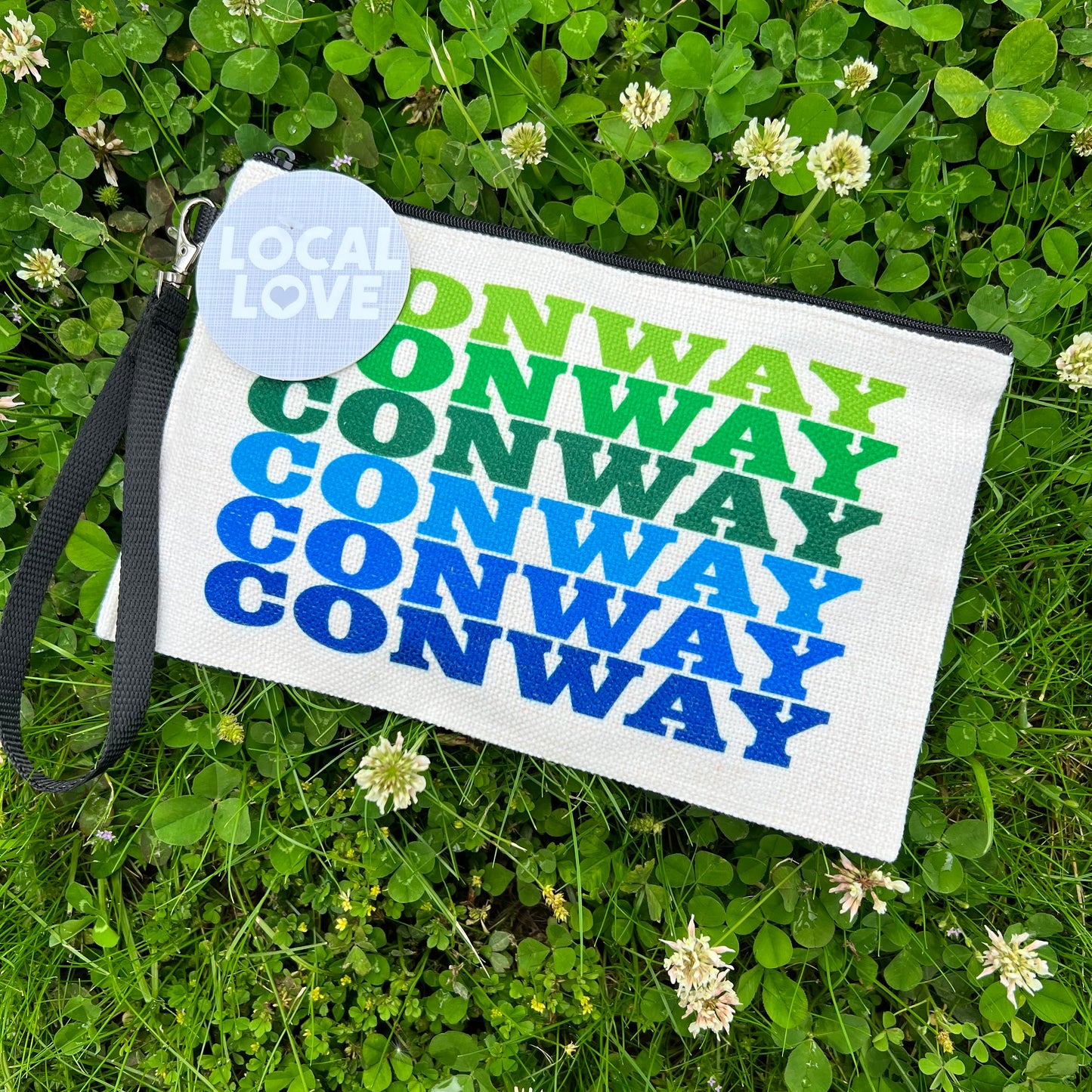 off-white zipper bag with "conway" printed in greens and blues colors laying in a field of clovers.