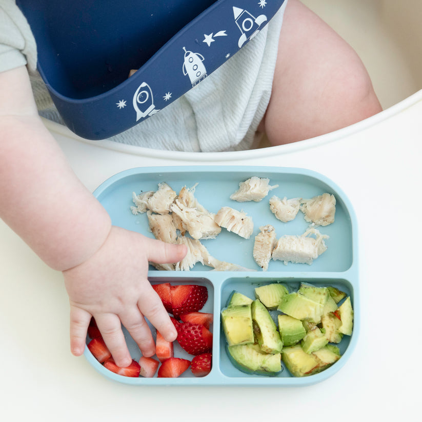 top view of a baby's hand reaching for fruits on a light blue silicone plate.