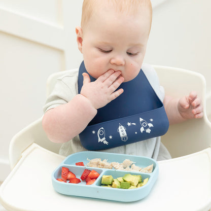 baby sitting in hairchair feeding themself from a blue silicone plate.