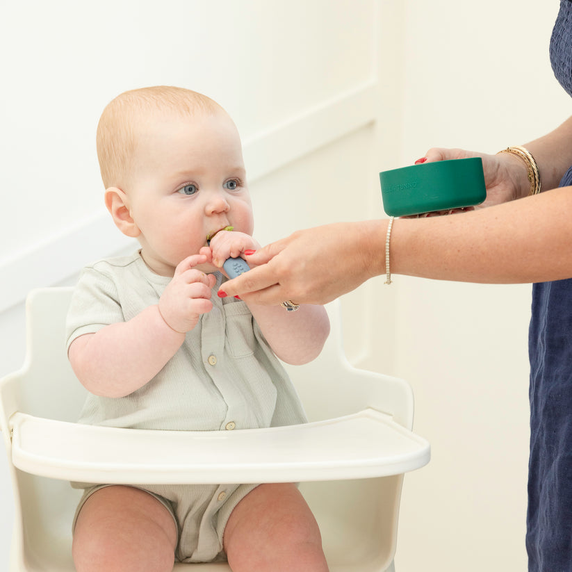 grown-up holding green silicone bowl and feeding baby sitting in a highchair.