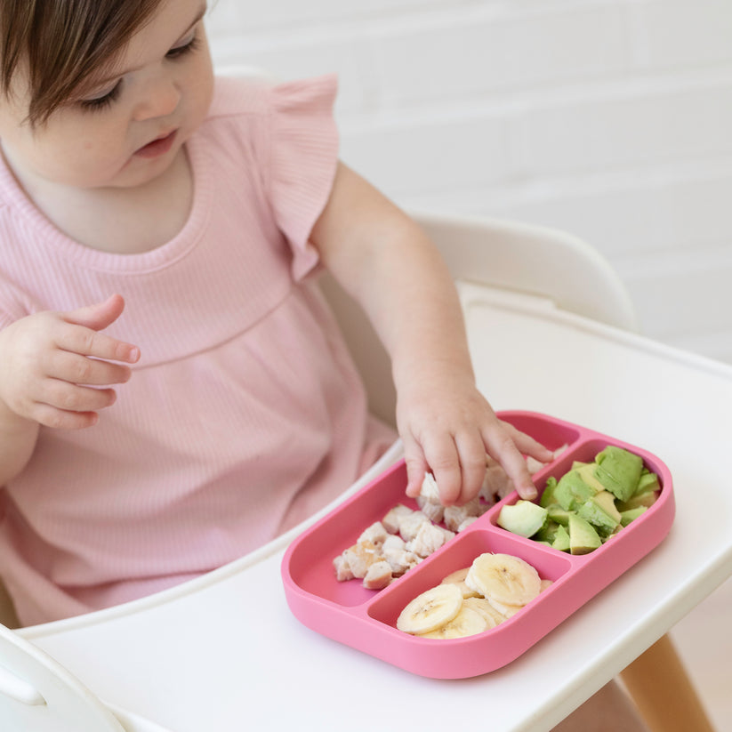 baby sitting in highchair feeding themself from a pink silicone plate.