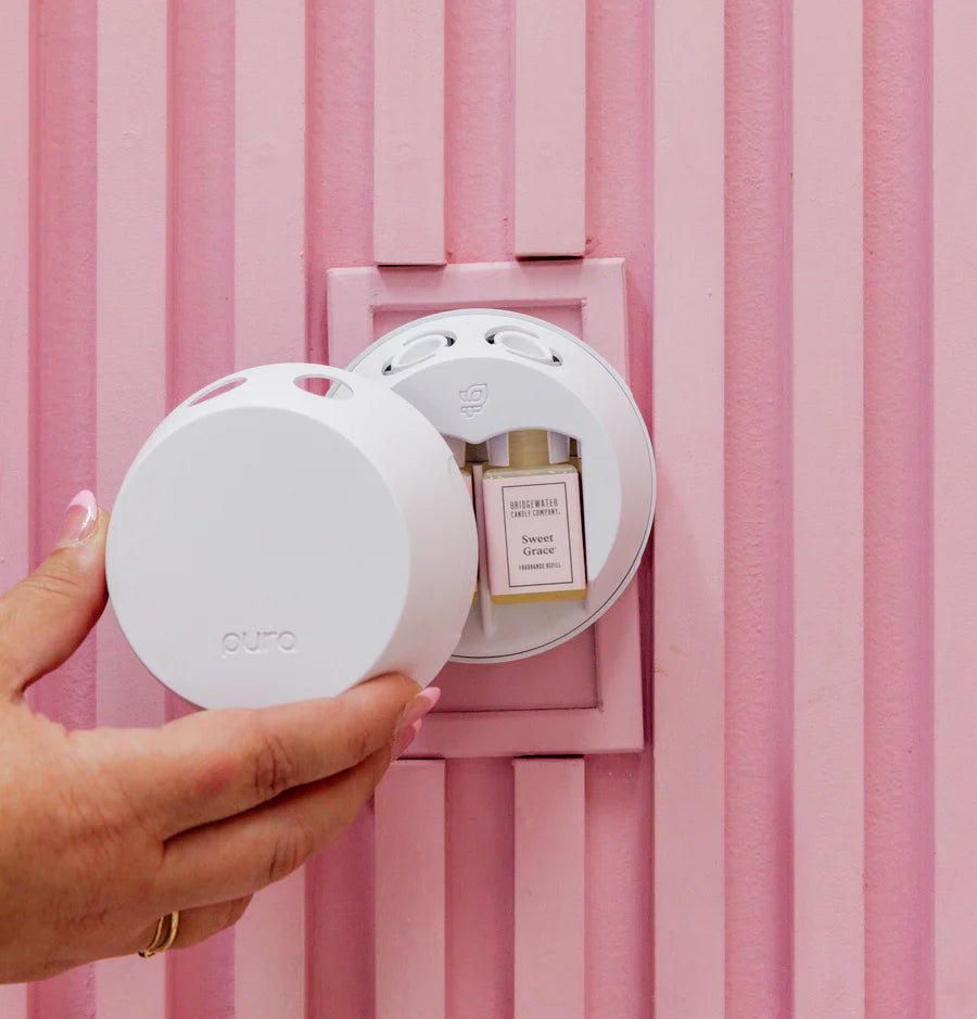 hand holding cover to pura device plugged into pink stripe wall filled with  vials of sweet grace