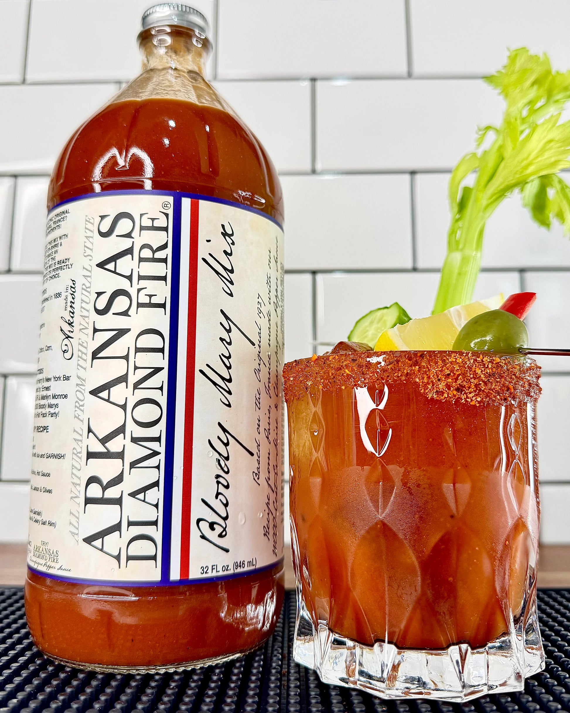 Bottle of Arkansas Diamond Fire Bloody Mary mix next to a glass with a bloody mary and celery stem in it on a white tiled background