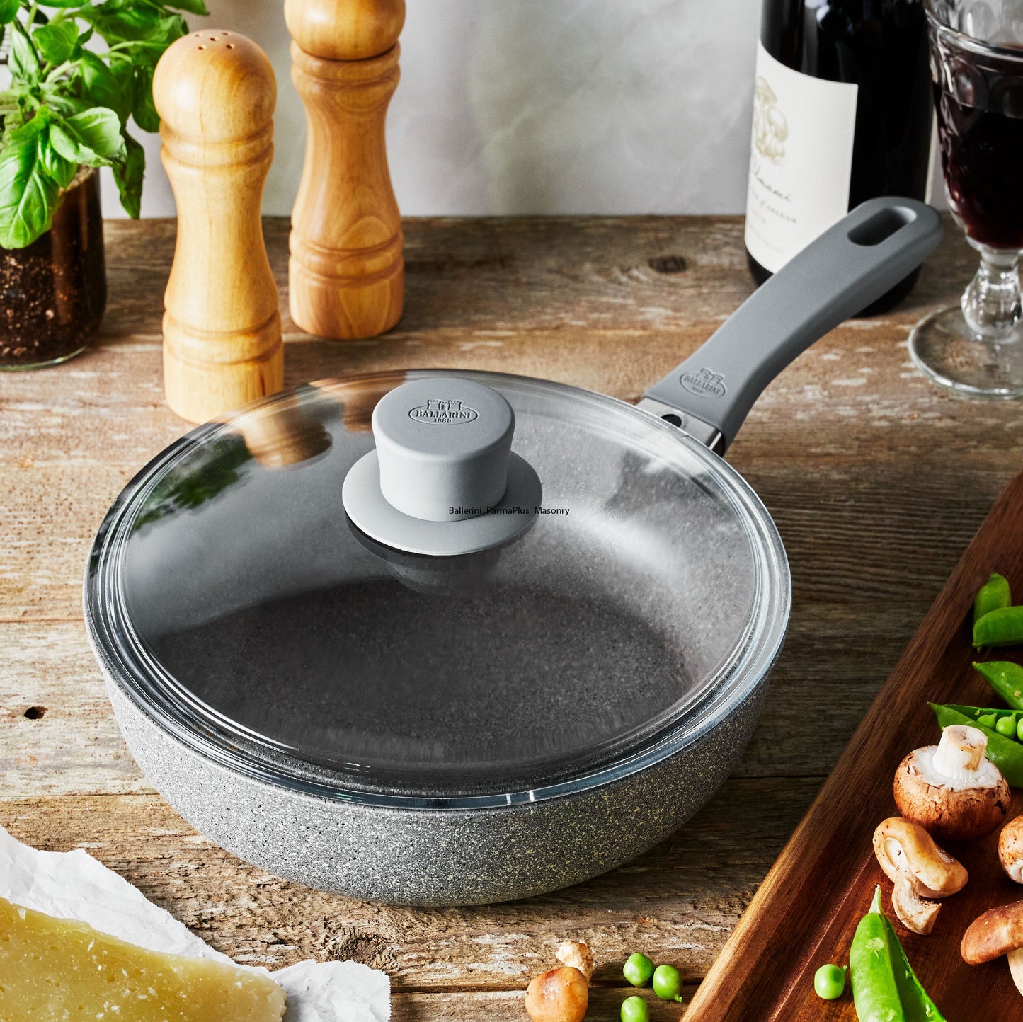 saute pan with lid set on a wooden table with veggies, wine bottles, and seasoning around it.