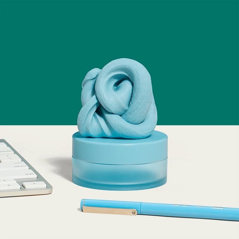  light blue fidget putty with a twisted, looped shape resting on a matching round translucent base, styled on a white surface with a teal background, part of a keyboard at left, and a blue pen at bottom right