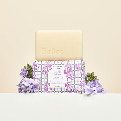 bar of lilac dream soap stacked on another bar wrapped in paper packaging with lilac flowers arranged around it.