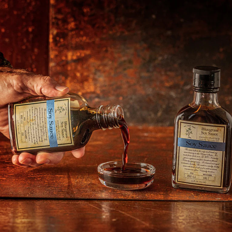 Hand pouring soy sauce from a bottle into a small glass bowl on a wooden surface.