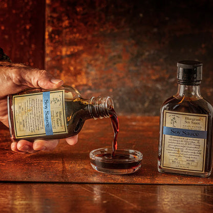 Hand pouring soy sauce from a bottle into a small glass bowl on a wooden surface.