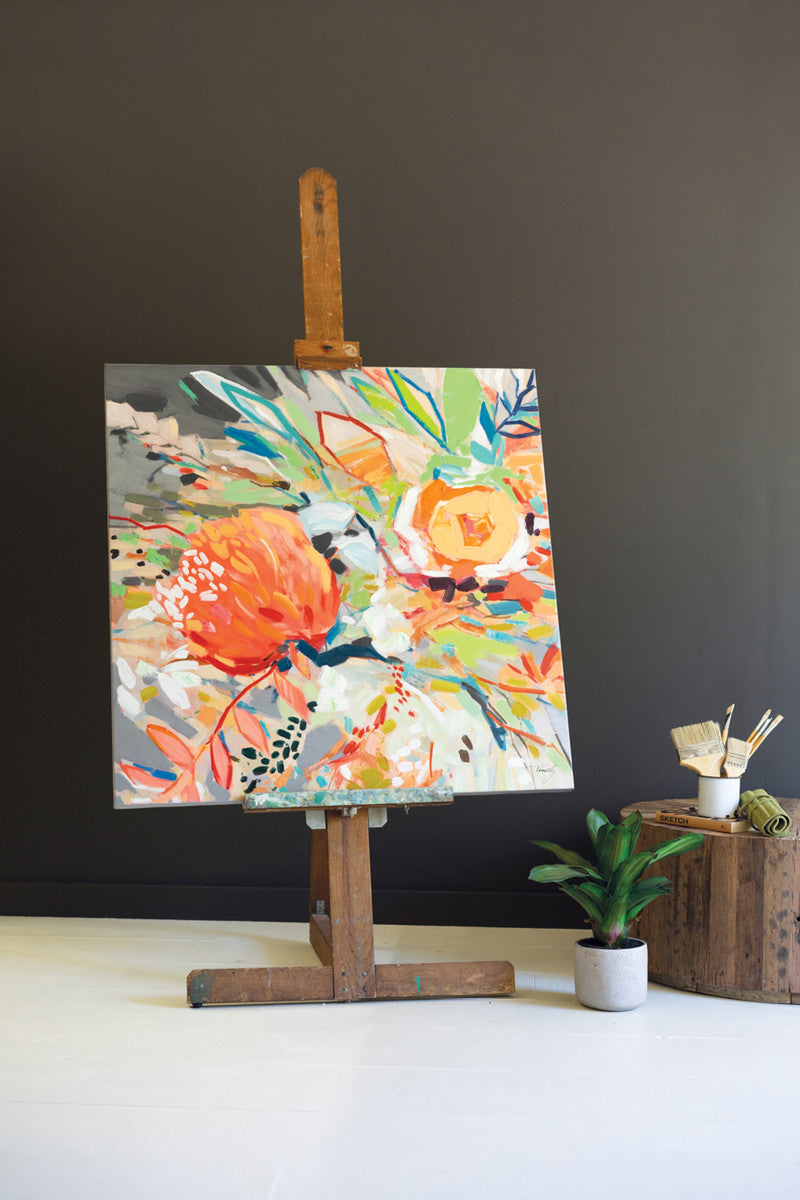 floral art in wooden easel set on a table with a plant and paint brushes.
