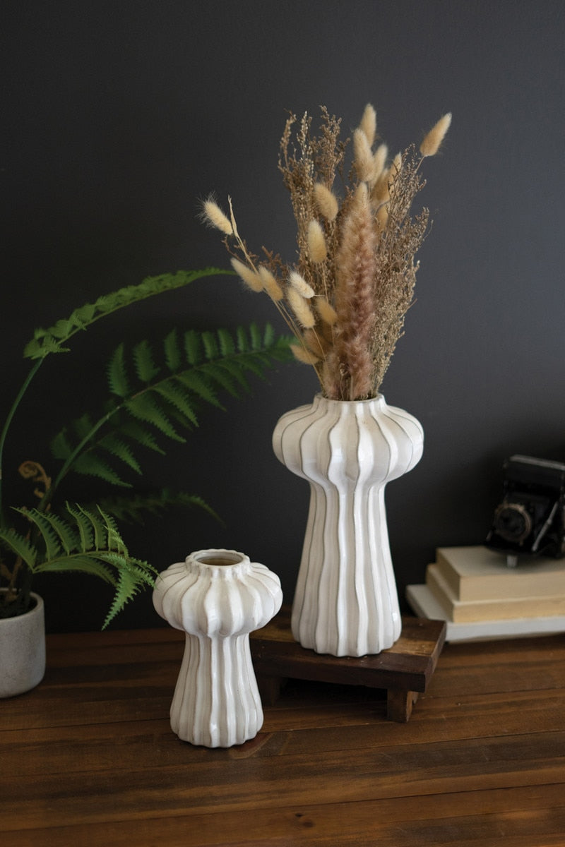 White textured vases with dried stems in it arranged on a wooden surface against a dark wall.