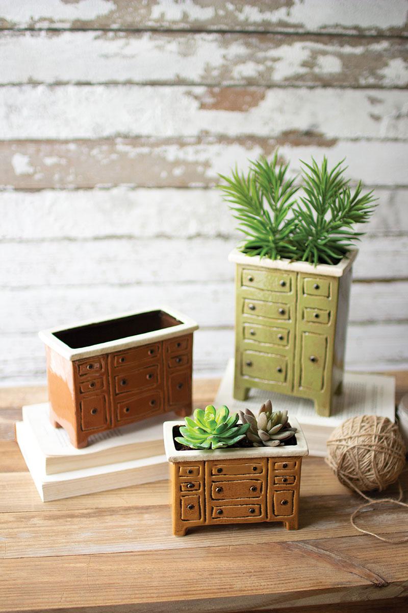 3 styles of ceramic chest of drawers planters, 2 are filled with succulents, all are arranged on a table with books and a ball of twine.