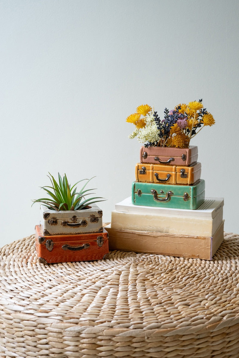 Two sizes of ceramic suitcase planters arranged with books on a rattan table.