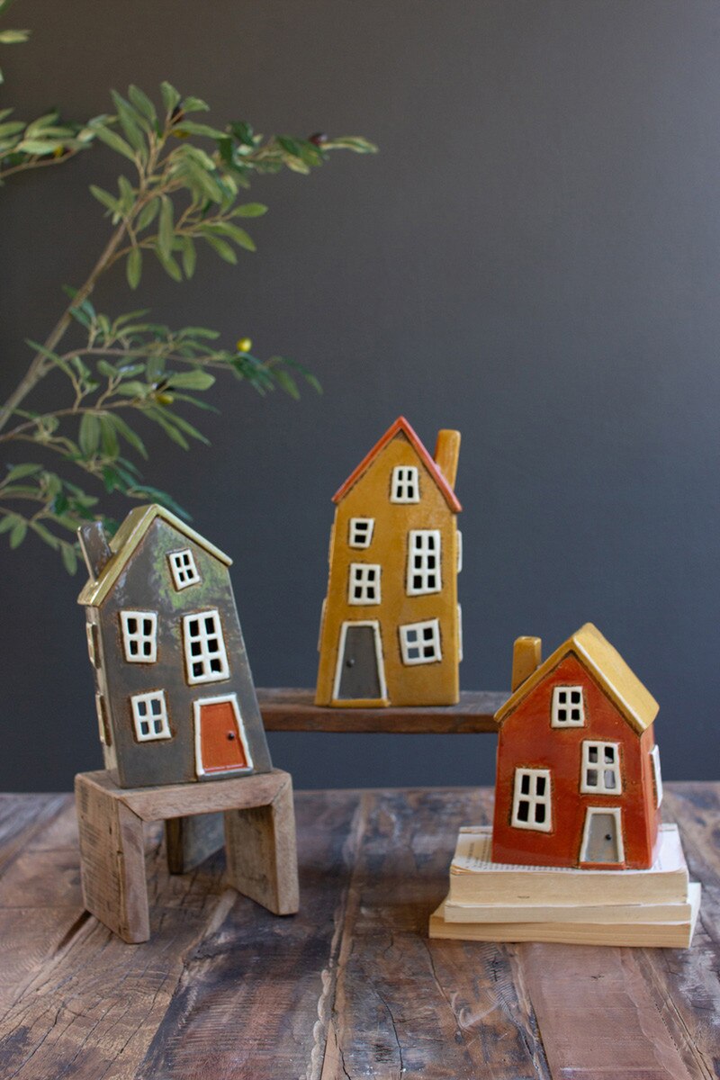 Three sizes and colors of ceramic wonky houses set on wooden risers and books.