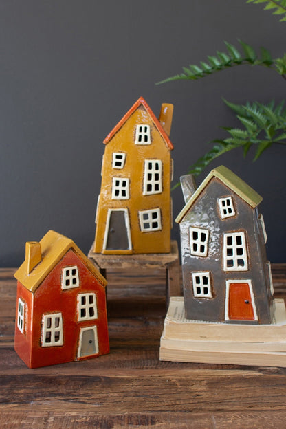 Three colors and sizes of ceramic wonky houses Arranged on a wooden table with books and a plant in the background.