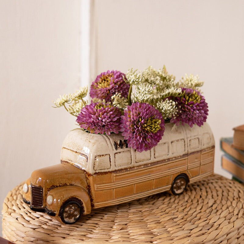 planter shaped like a classic yellow school bus filled with purple and white flowers set on a woven stool
