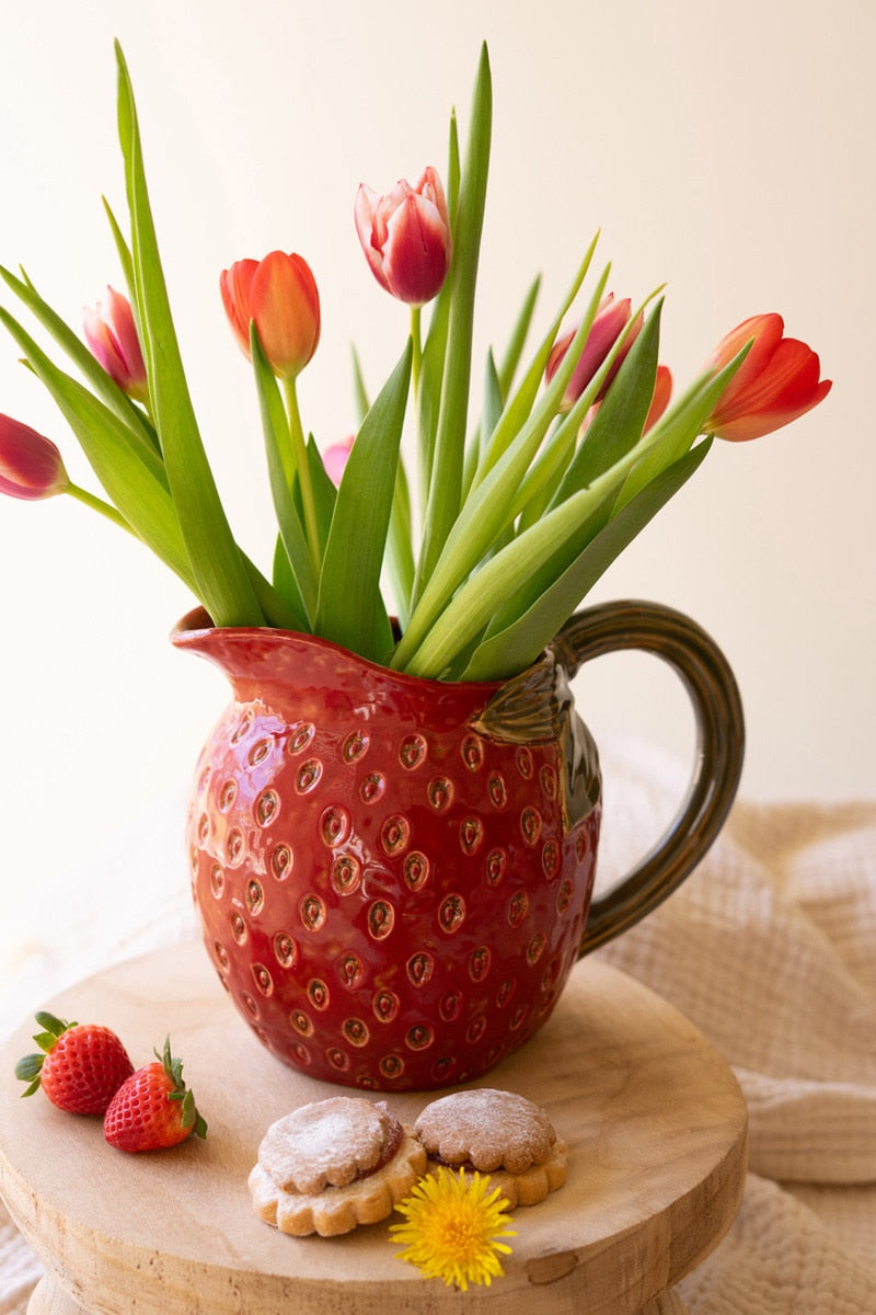 Red strawberry shaped pitcher filled with tulips set on a wooden surface with cookies and strawberries.