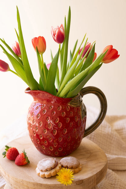 Red strawberry shaped pitcher filled with tulips set on a wooden surface with cookies and strawberries.
