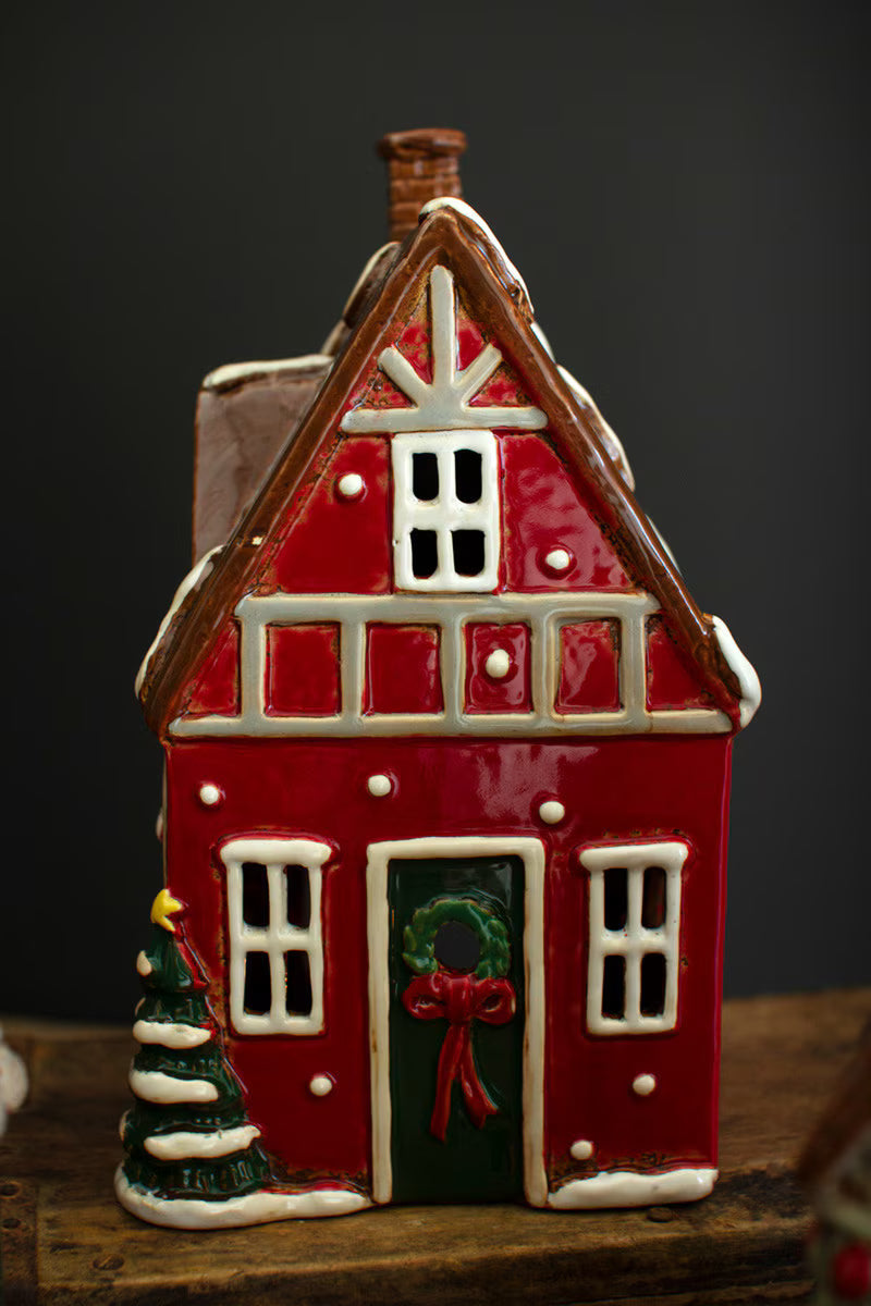 A red ceramic farmhouse house figurine with white trim, a green door with a red bow and wreath, and a small green Christmas tree beside it, set on a wooden surface against a dark background.
