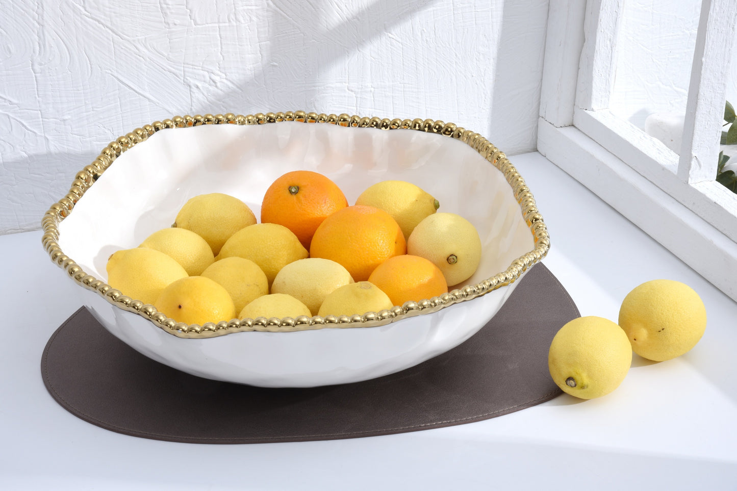 White bowl with gold rim containing oranges and lemons on a white surface.