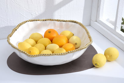 White bowl with gold rim containing oranges and lemons on a white surface.