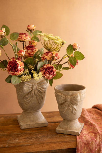 Two sizes of decorative urns with bow design on them, larger one has flowers in it shown on a wooden surface.