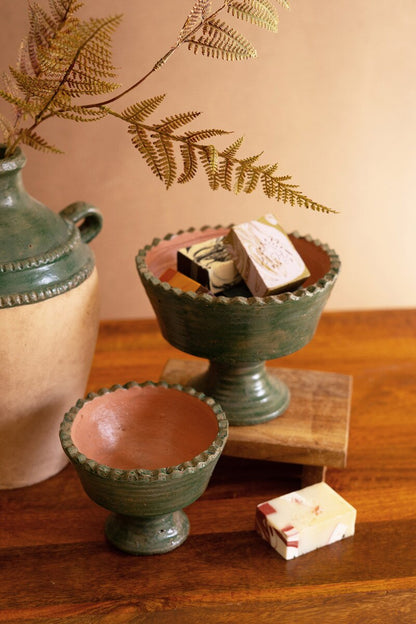 Two green ceramic footed bowls with soap in them on a wooden surface, with a vase and ferns in the background.