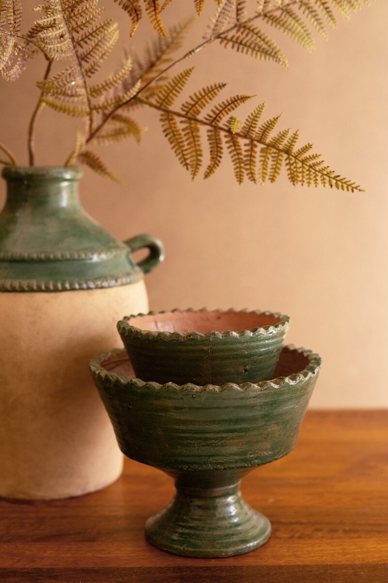 Green ceramic compotes stacked on a wooden surface with a beige wall and plant in the background