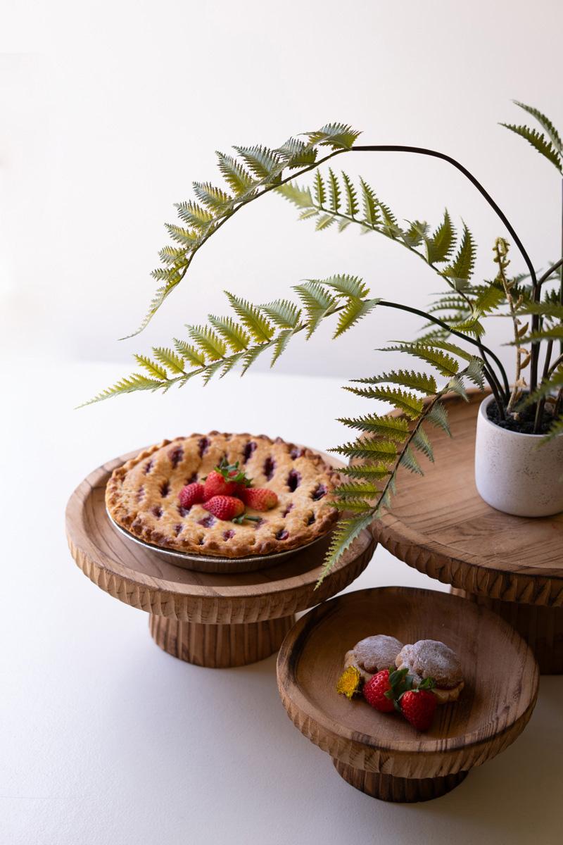 Wooden cake stands with a pie and dessert on a white surface with a plant in the background