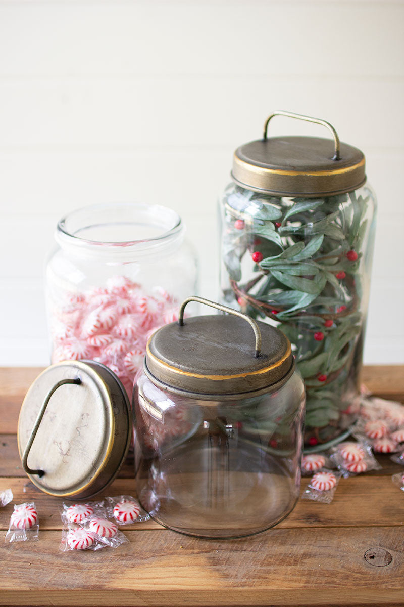 3 sizes of glass canisters on a wooden table, large is filled with greenery and red berries, medium has lid off and is filled with peppermints, small is empty.