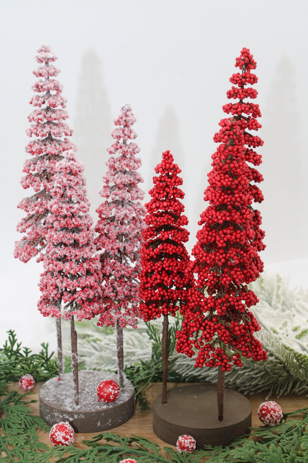 3 red berry trees and 3 frosted berry trees on bases arranged with greenery on a round wooden base arranged on a table with greenery