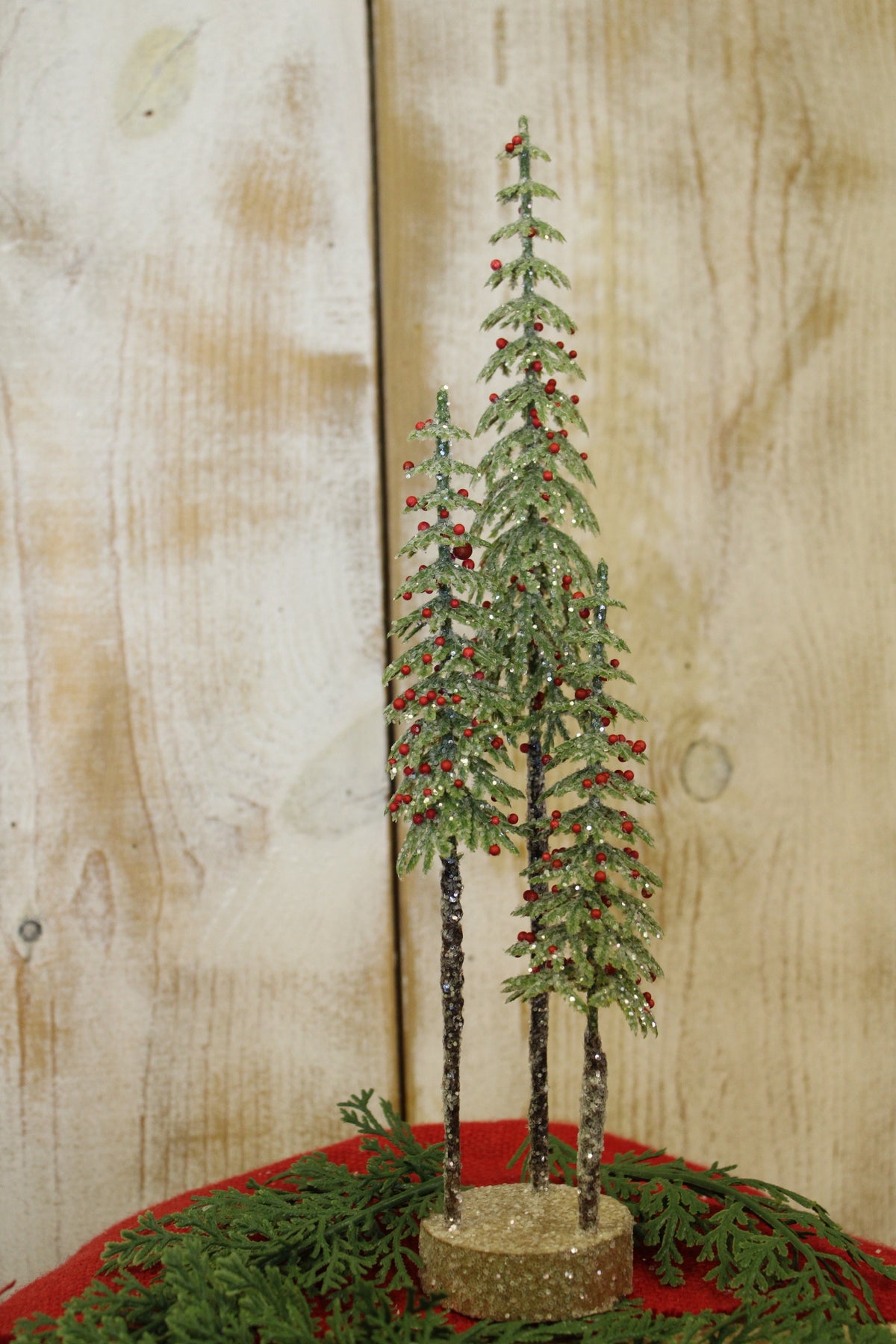 three slim green glitter frosted trees with red berries sharing a log slice wood base on decorated surface