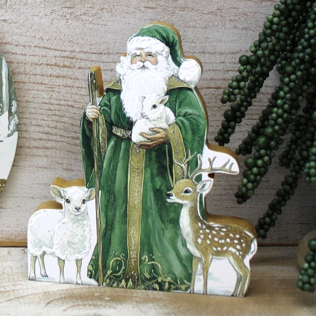 A decorative Santa Claus figurine wearing a green robe and holding a white lamb, with a deer and another lamb in front, displayed on a wooden surface against a wood-grain background.