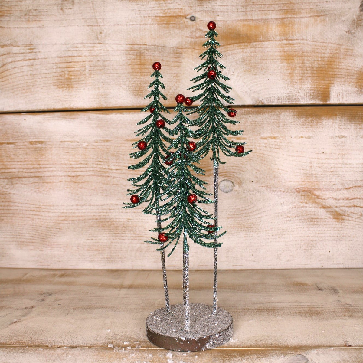three glitter coated faux trees with red ornaments sharing a wood base against an wood background