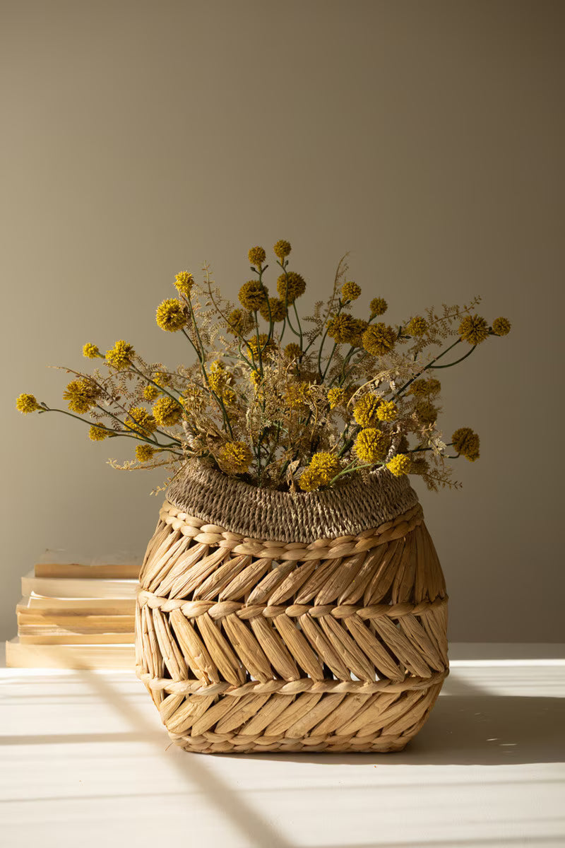 A woven natural-fiber basket vase filled with yellow dried flowers, set on a white surface against a warm beige background with soft window-light shadows.