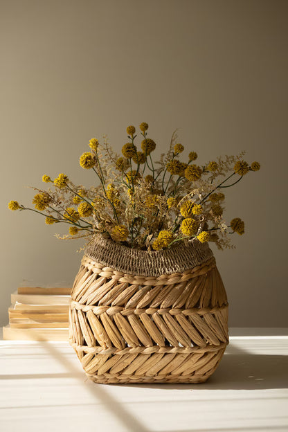 A woven natural-fiber basket vase filled with yellow dried flowers, set on a white surface against a warm beige background with soft window-light shadows.