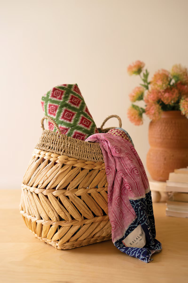 A woven hyacinth basket sits on a light wood table, holding a folded multicolor patterned blankets and draped a pink patterned scarf with a navy blue border. A terracotta vase with pink flowers and stacked books are softly blurred in the background.