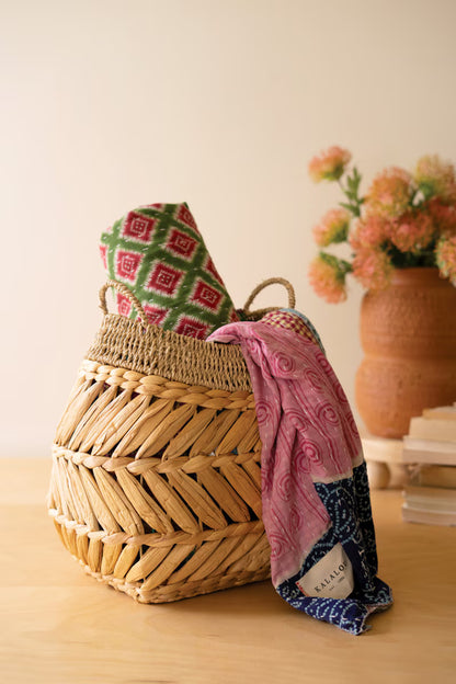 A woven hyacinth basket sits on a light wood table, holding a folded multicolor patterned blankets and draped a pink patterned scarf with a navy blue border. A terracotta vase with pink flowers and stacked books are softly blurred in the background.