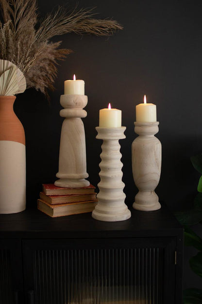 3 styles of wooden candle holders arranged on a side table with books, a vase, and lit candles on them.